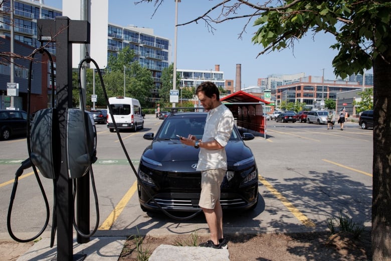 Man in front of his electric vehicle while its plugged into a charger in the parking lot of a  grocery store.