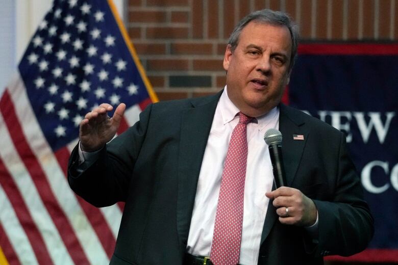 Grey haired man with red tie speaks into microphone in front of an American flag