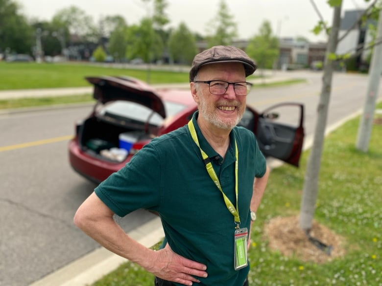 Garnet wears a dark green shirt with a light green lanyard. He stands in front of his red car filled with meals to be delivered. 