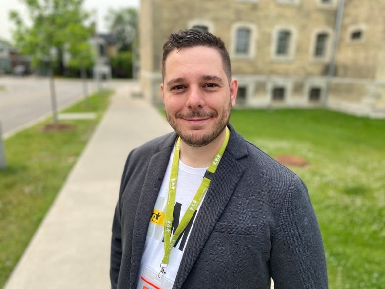 Adriano Murarotto wears a white t-shirt and a grey blazer and a green lanyard. He is smiling while standing on the sidewalk. 