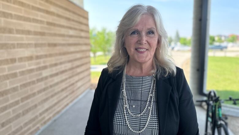 An older woman looks at the camera and smiles. She's standing outside a building with grass and trees in the back. She wears a pearl necklace and black blazer over a checkered shirt.