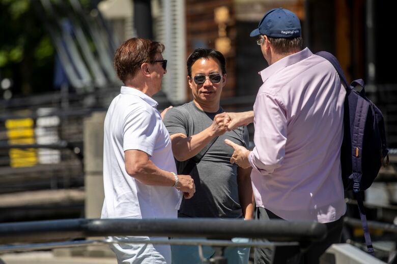 A man in sunglasses bumps fists with another man in a baseball cap while a third man looks on.