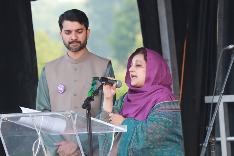 A dark-haired man with a neatly trimmed beard and moustache looks on as a woman wearing a magenta headscarf speaks into a microphone.  