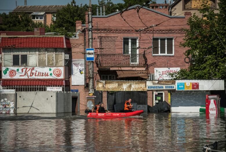 Three people are shown on a boat patrolling a town that is flooded, with small businesses shown in the background.