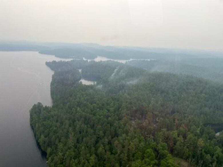 Smoke rises from a forest next to a lake on a smoky day overall.