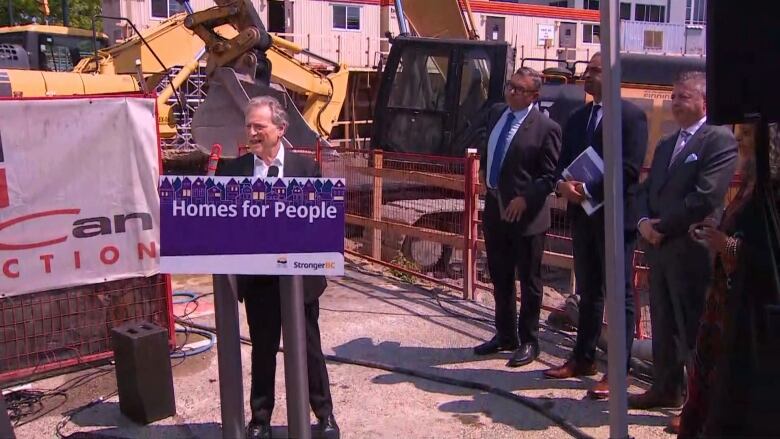 A man stands at a rostrum in front of a construction site with other politicians and a developer.