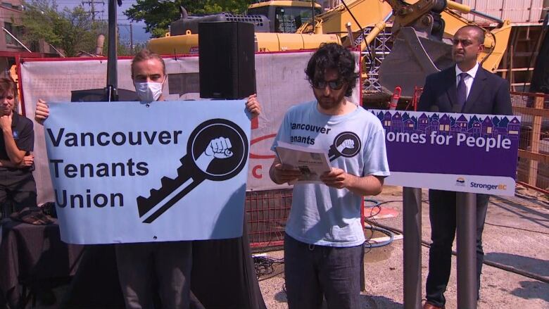 Two men in the foreground wear blue Vancouver Tenants Union T-shirts. One holds up a blue Vancouver Tenants Union sign. Ravi Kahlon stands behind a rostrum staring at the two men.