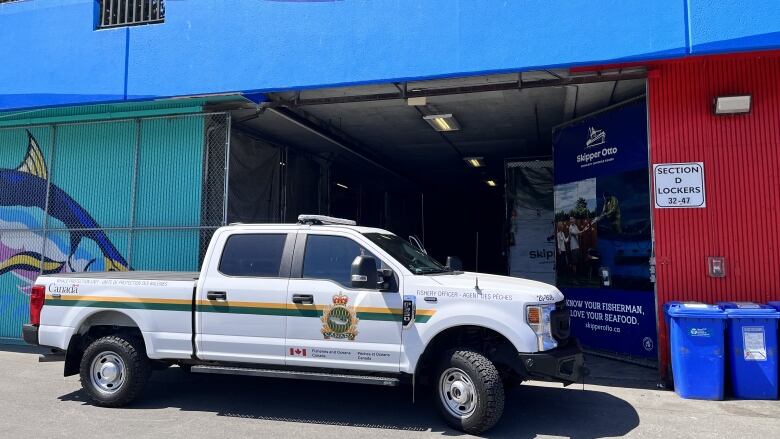 A pickup truck with Fisheries and Oceans markings is parked beside the open doors of a red and blue warehouse that has a large cartoon fish on the blue part of the wall.