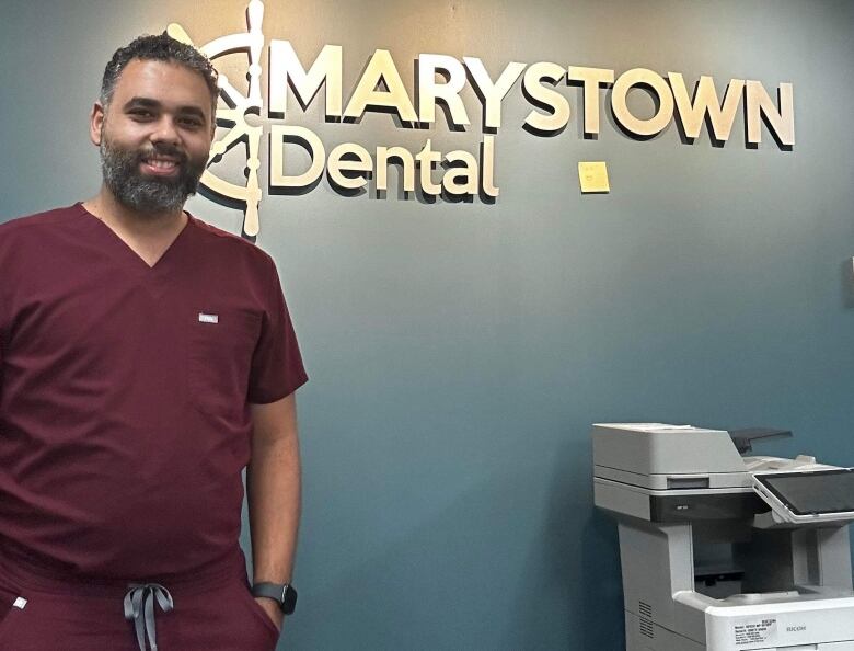 A man in burgundy medical scrubs poses in front of a wall with a sign that reads 'Marystown Dental.'