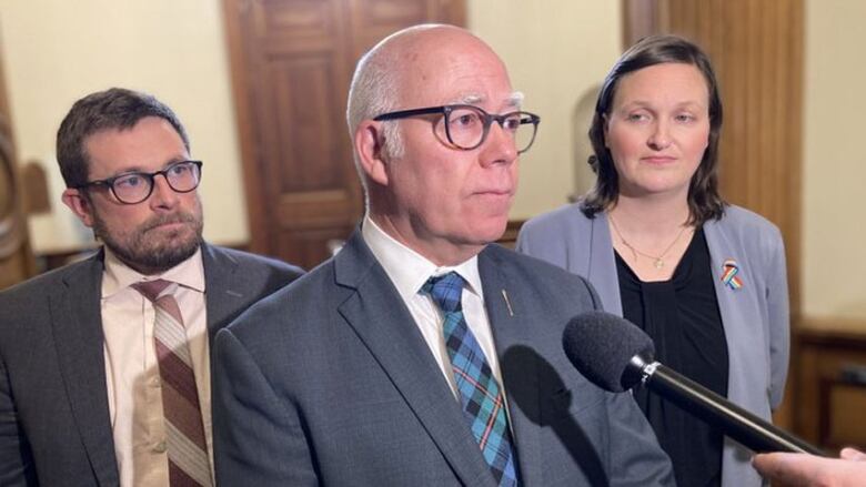 A man with grey hair and glasses, wearing a grey suit, flanked by another man and a woman, speaks into a microphone.