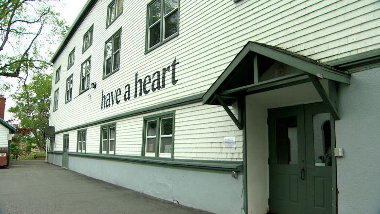 A white wooden-clad building with green trim and a door has black lettering stencilled on the side reading 