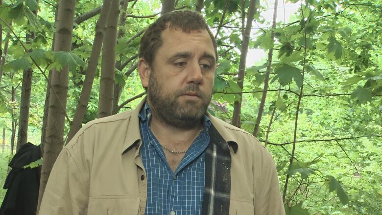 A white man with a tan jacket and blue-checked shirt stands in a wooded area with green foliage around him