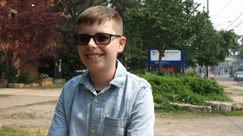 A kid wearing sunglasses sits in front of the Earl Kitchener school sign and smiles.