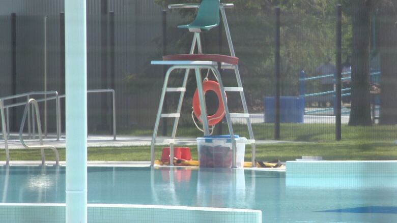 A public pool is full of water, but nobody is swimming. A lifeguard chair towers over the edge. A life preserver hangs from the back. Underneath the chair is a container filled with first aid packs.