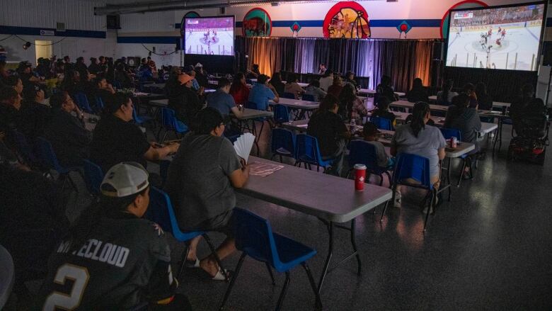 People gather in a large hall to watch a hockey game on two screens.