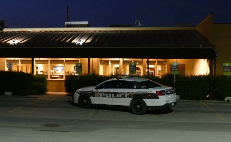 A black and white police cruiser sits parked in front of the windows of a restaurant at night.