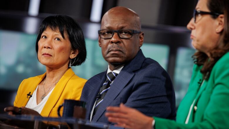 A woman in a yellow jacket sits beside a man in a dark suit. Booth look deep in thought.