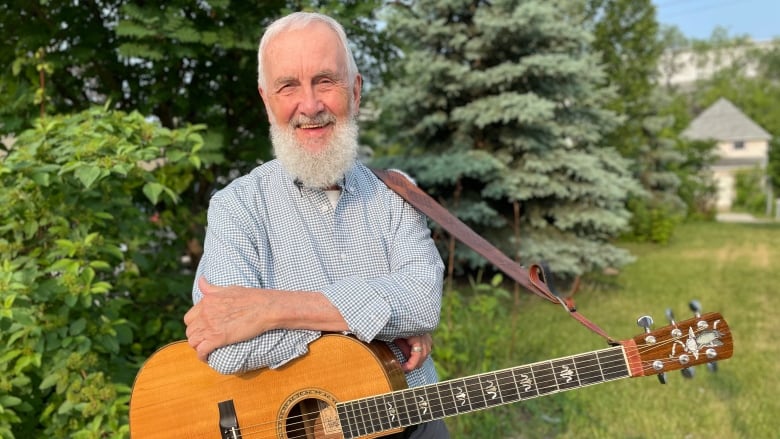 A man white short white hair and a white beard, wears a blue and white checkered shirt and holds a guitar while standing outside on a green lawn.