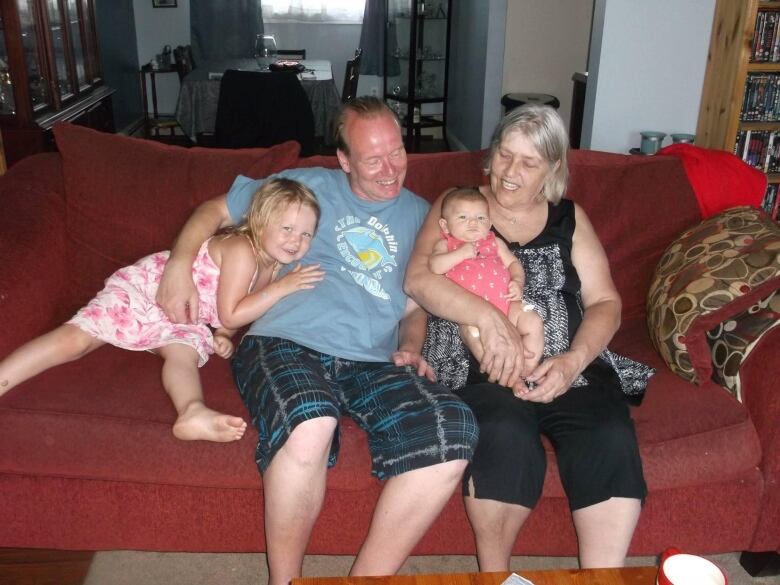 An older man and woman sit on a red couch with their grandchildren. One is a young girl with blonde hair and the other is a baby. 