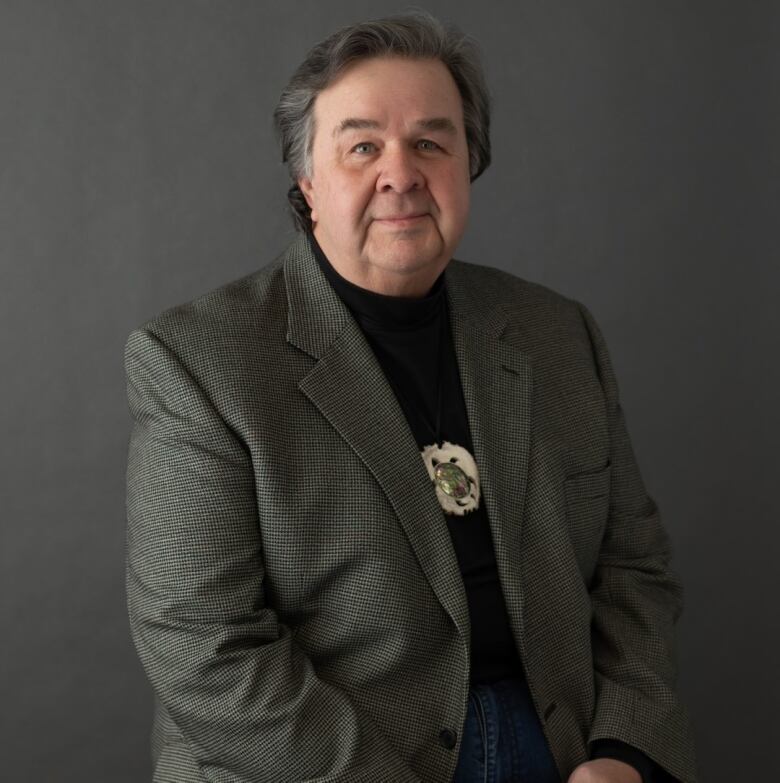 An Indigenous man poses with a grey blazer, black shirt and a turtle medallion. 