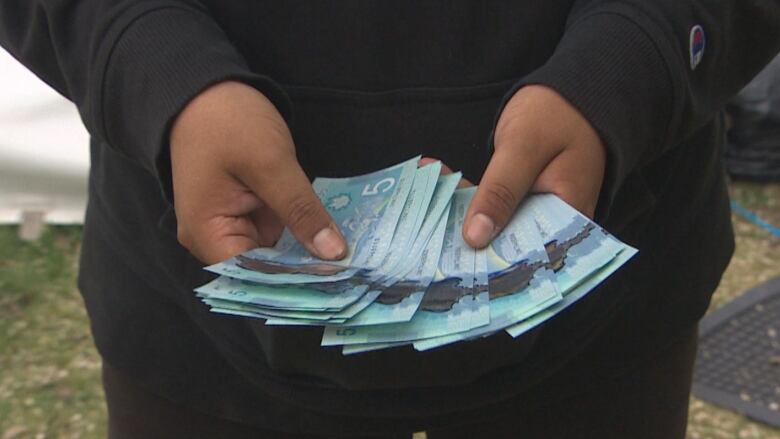 Two hands are pictured showing off a stack of five dollar bills to the camera.