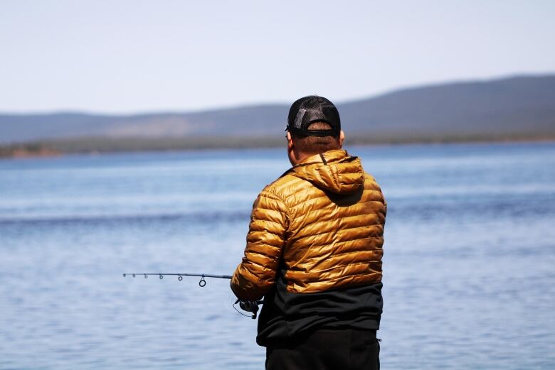 A man in a yellow jacket stands looking out towards mountains while fishing on a lake. 