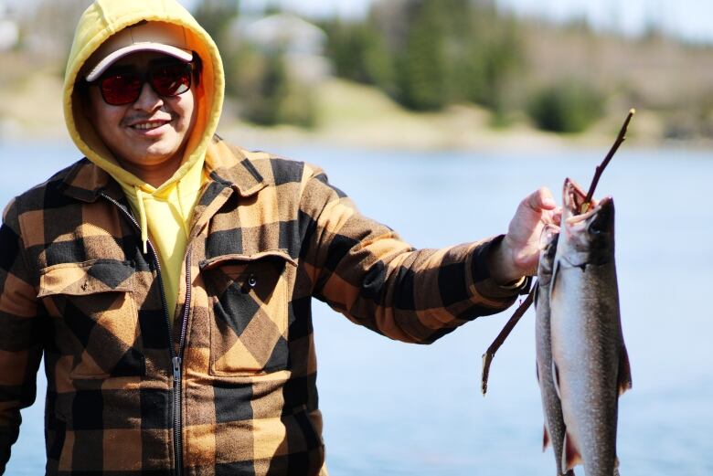 A man in a plaid jacket holds up three fish with a stick through the gills. 