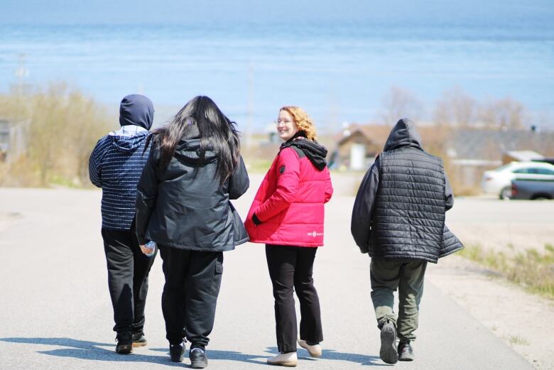 A group of people walk down a street with the ocean in the background. One is turned around and smiling. 