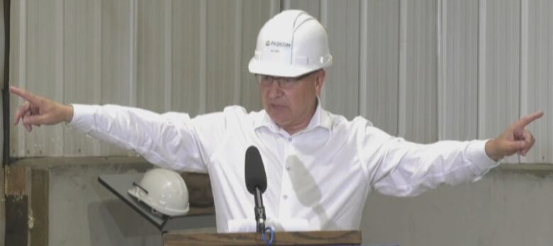 A man in a white shirt and white hard hat stretches his arms wide as he speaks into a microphone.