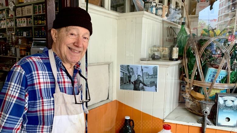 A man in a hat and apron stands outside a shop, in the background are tins of Italian food.