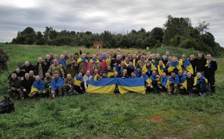 A group of dozens of men, some holding blue and yellow Ukrainian flags, in a field.