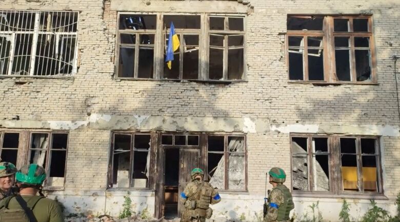 People in camouflage uniforms stand in front of a damaged building with a blue and yellow flag hanging from a damaged window on the upper level. 