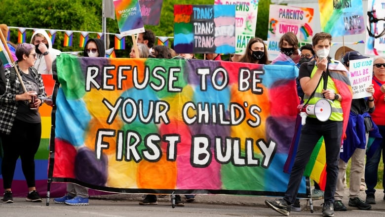 People at a protest carry a large multicoloured sign saying 'Refuse to be your child's first bully.'