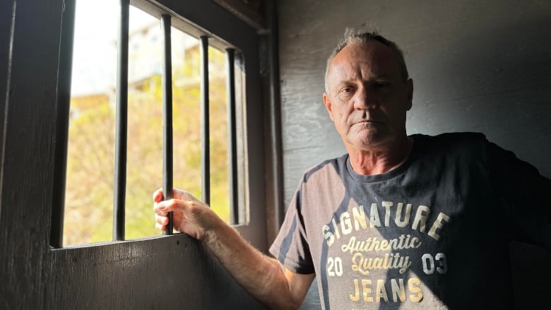 A man stands inside a makeshift jail cell, with one hand wrapped around the bars on the window.