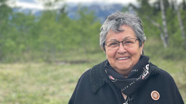 A woman with short grey-black hair, wearing a black jacket and glasses, smiles as she poses for a portrait outside.