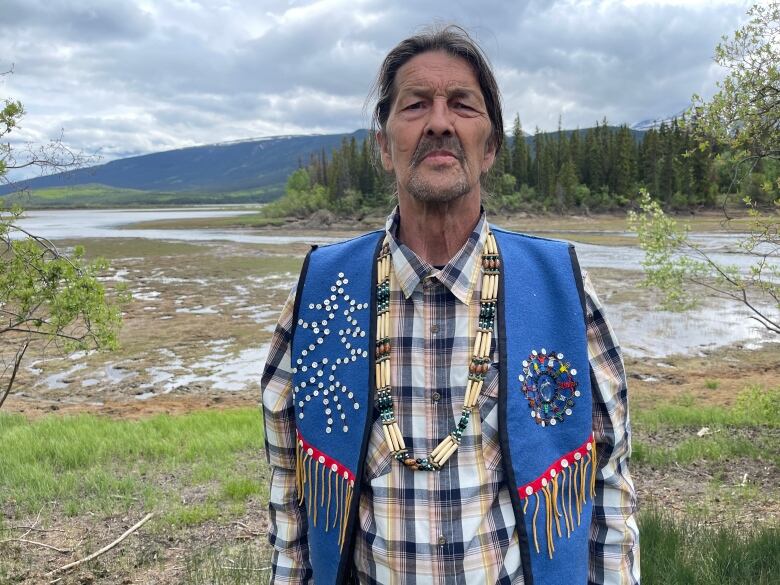 An Indigenous man with a mustache and long hair pulled into a ponytail poses for a portrait outdoors. He is wearing a plaid shirt, a beaded blue vest and a wooden bead necklace.