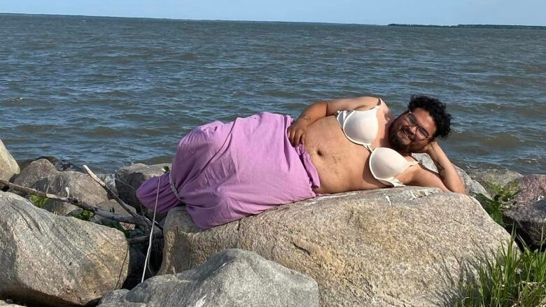 Julian Baptiste laying on a rock on the Eagle Lake Island beach with a white bra representing Ebb and Flow First Nation