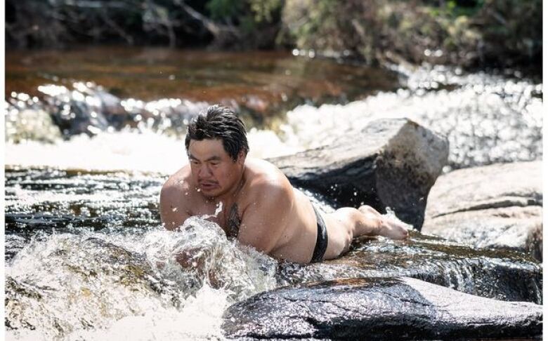 A man in a swimsuit lies stomach-down on a rock with rushing water around him.