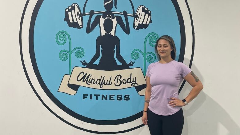 A woman poses in front of a sign in a gym that reads Mindful Body Fitness. 