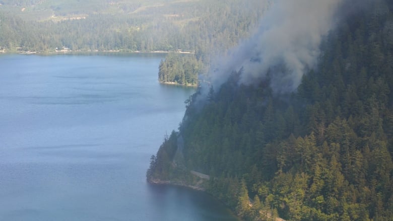 a fire burns with smoke above a lake-side highway, with a helicopter at the bottom carrying water.