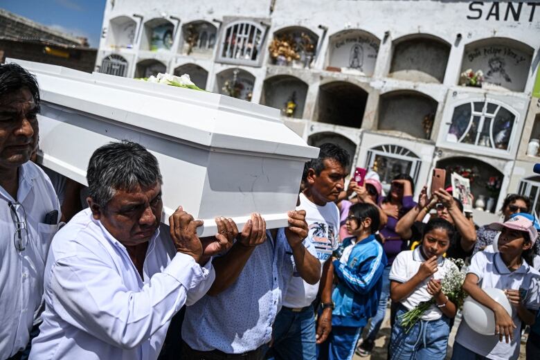 Men carry a white coffin on their shoulders past a crowd of people in a cemetery.