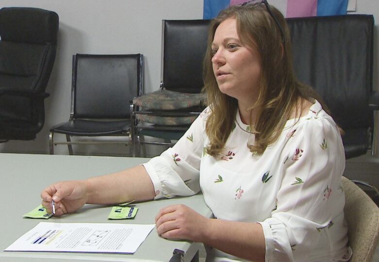 A woman sits at a table holding a small narrow strip of paper. There are two green packages holding drug test kits in front of her. 