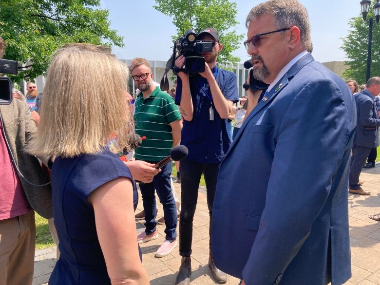 A woman with blond hair and a tall man in a blue suit face each other outside while TV cameras look on.