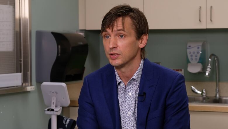 Dr. Jared Van Bussel sits in a clinic exam room, with a sink and portable blood pressure machine behind him. He's wearing a blue suit and looking off camera.