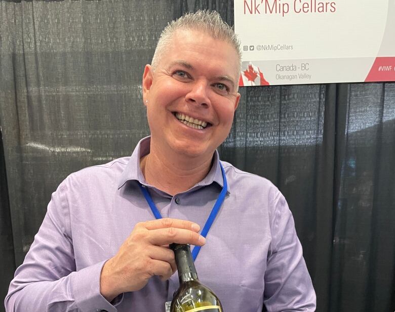 A man in a violet dress shirt holds a bottle of wine, smiling.