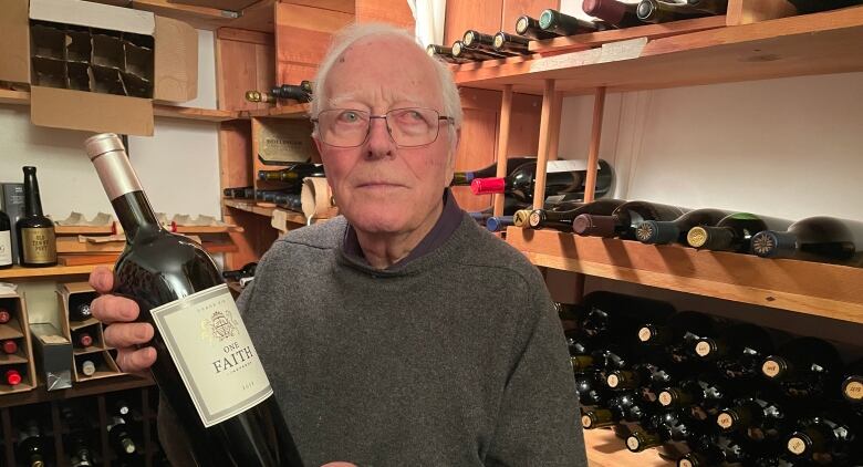 An older man holds a bottle of wine in front of a display of wines. 