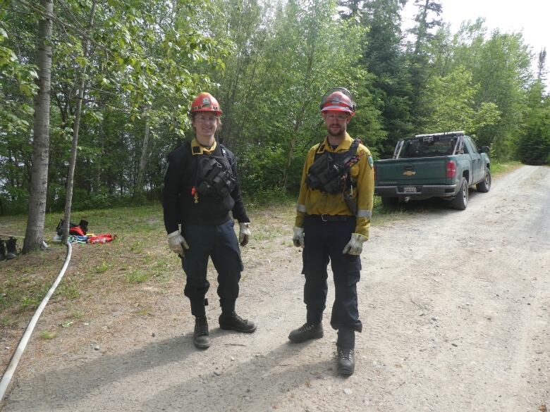 Two men in firefighting gear pose, smiling, on a road in a forest.