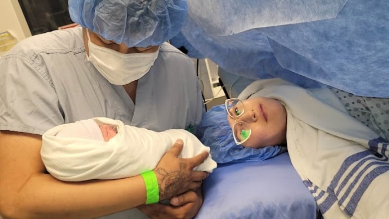 Catherine is in an operating room draped in surgical blankets. Her husband holds their new baby.