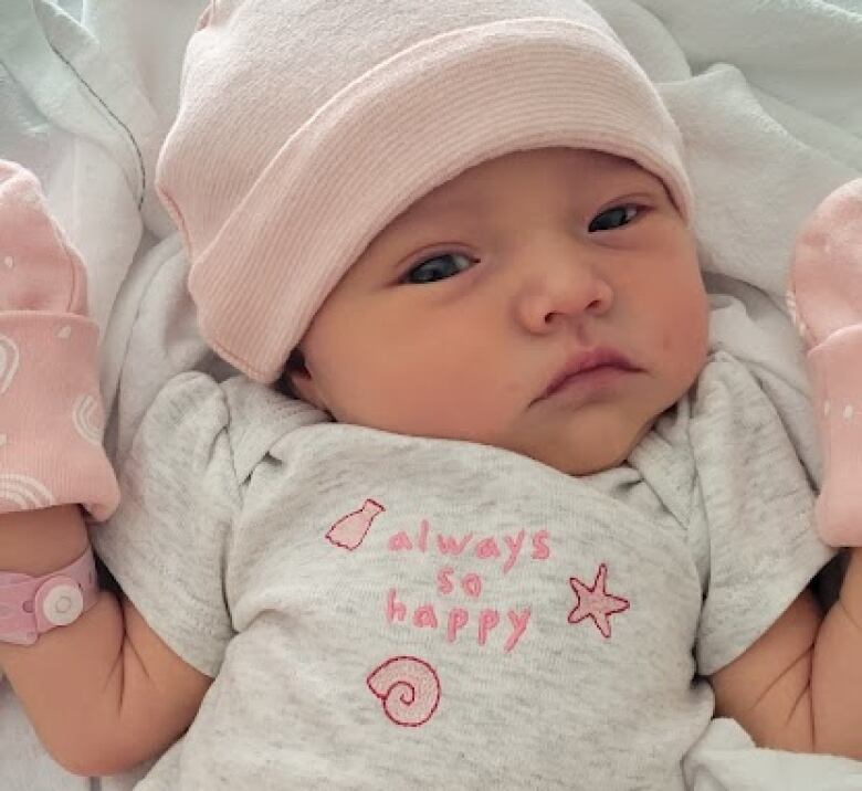 A Baby girl is dressed in pink and white wearing a pink toque and mitts.