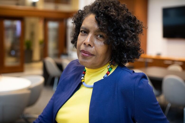 A Black woman with curly hair, wearing a yellow shirt and a blue coat, poses for a picture in a conference room.